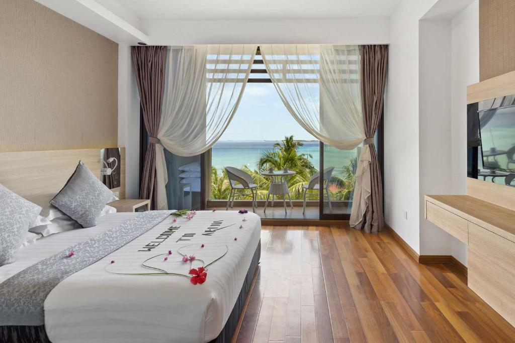 Modern hotel room with a neatly made bed, wood flooring, and a balcony with a sea view and palm trees overlooking bikini beach in maafushi